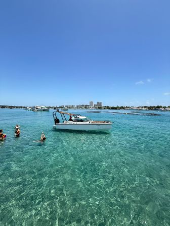 Sunny coastal scene with a white motorboat anchored in crystal-clear turquoise water, swimmers nearby and a distant city skyline under a clear blue sky.