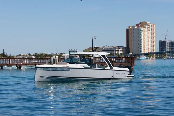 White motorboat cruising past a wooden pier on calm blue water in a sunny coastal marina, with waterfront high-rise buildings and a bridge in the background.