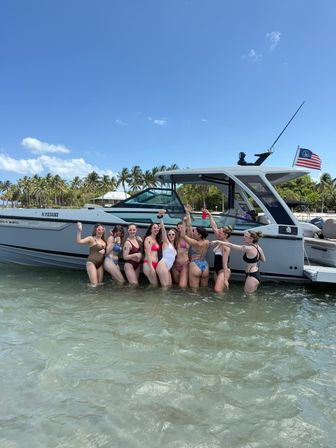 Group of friends in colorful swimsuits waist-deep in clear water beside a modern motorboat at a palm-lined Florida beach, toasting drinks under a bright blue sky with an American flag flying.