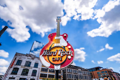 Giant guitar-shaped neon cafe sign with sunburst finish towering over a downtown music and entertainment district beneath a bright blue sky with fluffy clouds
