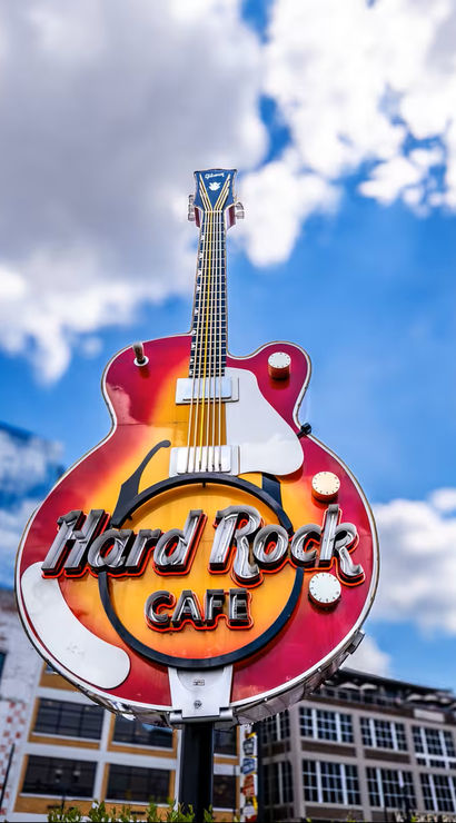 Giant guitar-shaped neon cafe sign with sunburst finish towering over a downtown music and entertainment district beneath a bright blue sky with fluffy clouds