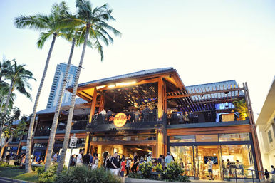 Evening scene of a two-story open-air wooden restaurant and bar with lit balcony, palm trees, and tourists strolling in a tropical coastal shopping district.