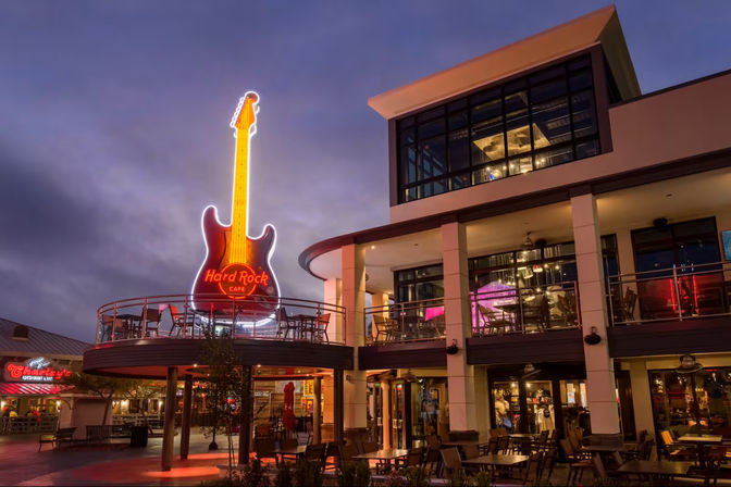 Giant neon guitar sign above a two-story restaurant with balcony patio at dusk in a lively waterfront entertainment plaza