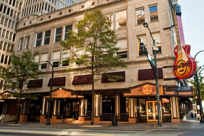 City corner restaurant in a stone multi-story building with trees, awnings and a giant red neon guitar sign marking a music-themed cafe.