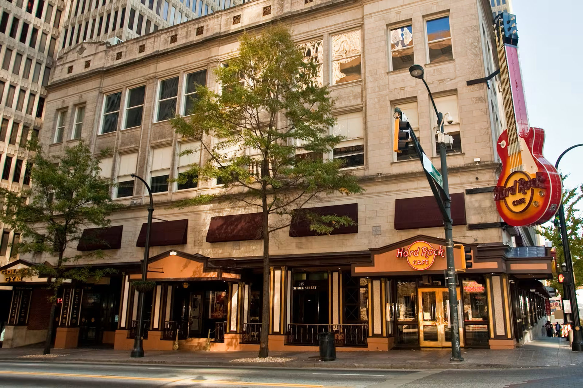 City corner restaurant in a stone multi-story building with trees, awnings and a giant red neon guitar sign marking a music-themed cafe.