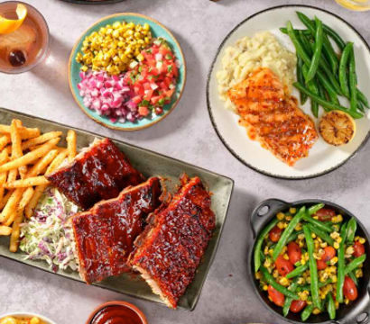 Vibrant tabletop spread of American-style dishes: glazed BBQ ribs with fries, grilled salmon with mashed potatoes and green beans, sliced steak, chicken salad and colorful corn-and-pico bowls.