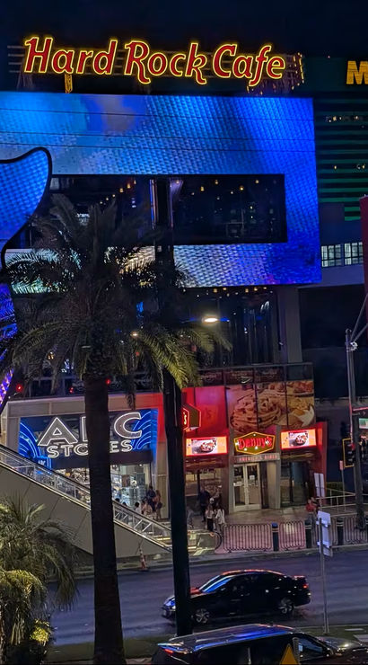 Vibrant nighttime Las Vegas Strip with a giant glowing blue guitar-shaped restaurant facade, palm trees, a large red soda mural on a neighboring building, hotel signage, and bustling traffic and pedestrians.