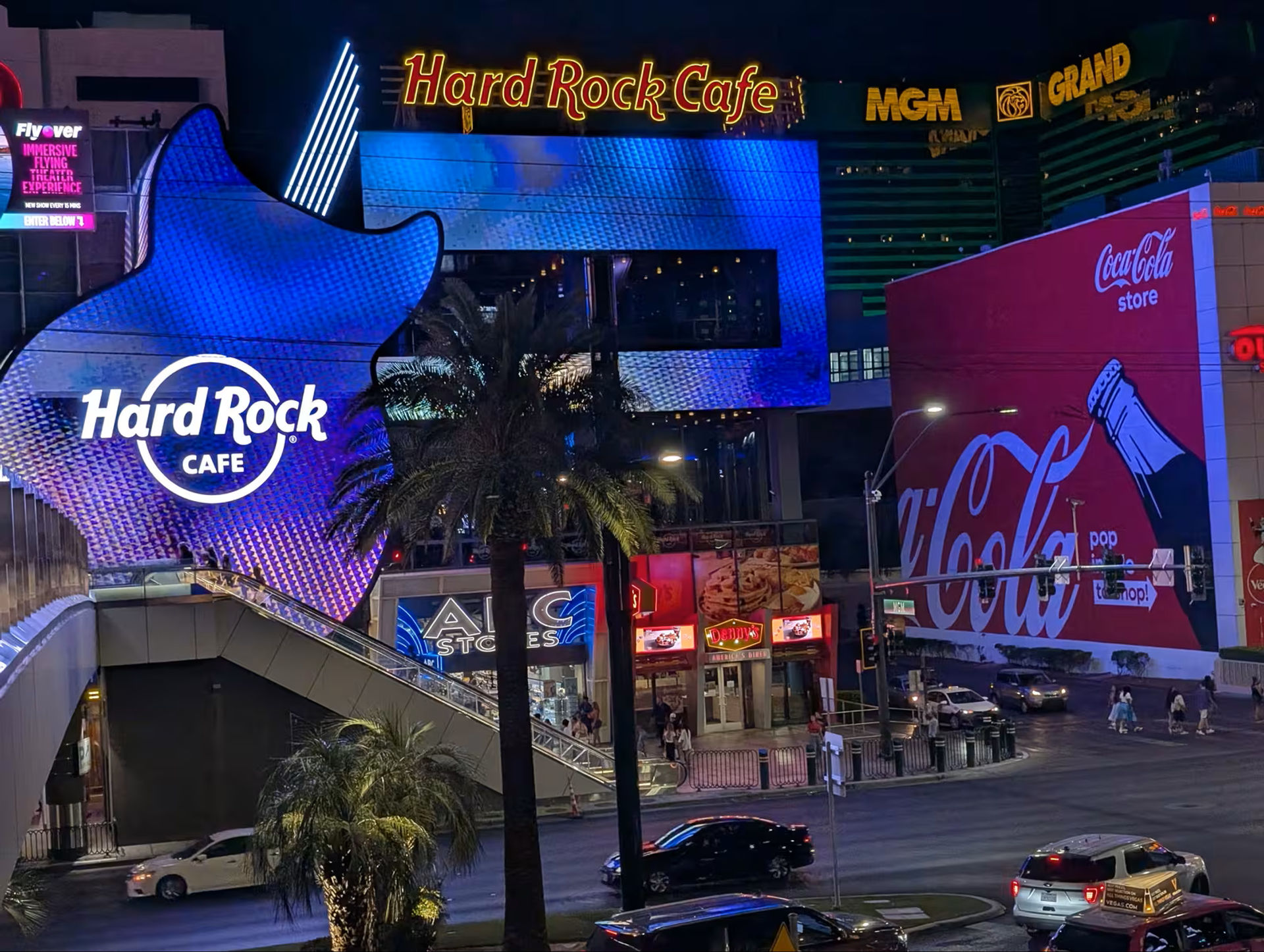 Vibrant nighttime Las Vegas Strip with a giant glowing blue guitar-shaped restaurant facade, palm trees, a large red soda mural on a neighboring building, hotel signage, and bustling traffic and pedestrians.