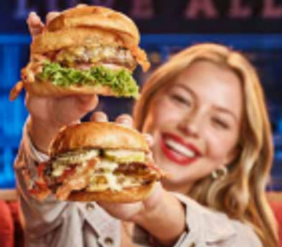 Smiling woman holding two stacked gourmet burgers toward camera in a colorful casual restaurant — juicy cheeseburgers with melted cheese, lettuce, pickles, and toasted brioche buns.