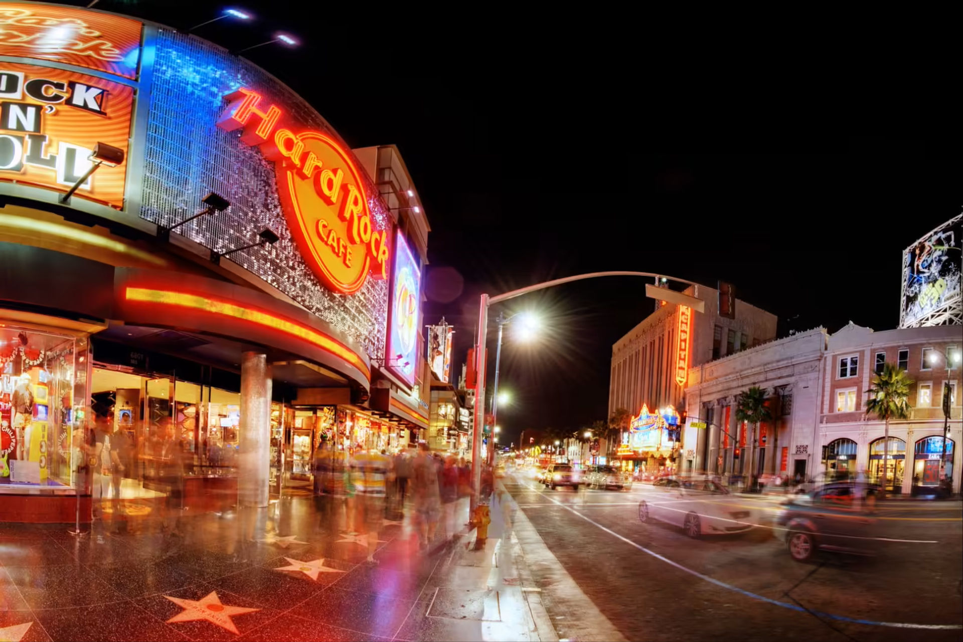 Nighttime Hollywood Boulevard scene on the Walk of Fame — bright neon rock-themed cafe sign and glittering marquee, stars on the sidewalk, bustling pedestrians and streaking cars.