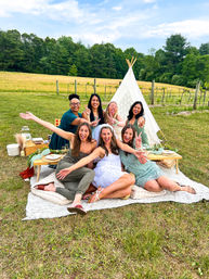 Seven friends enjoying a celebratory outdoor picnic on a blanket in a grassy meadow near vineyard rows, posing in front of a lace teepee with low wooden tables and rustic picnic decor, one person wearing a white dress and veil — cheerful group photo.