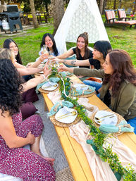 Group of friends toasting with champagne at a styled outdoor picnic in a grassy park — low wooden table with woven placemats, gold cutlery, pastel linens, leafy garland and lace teepee backdrop.