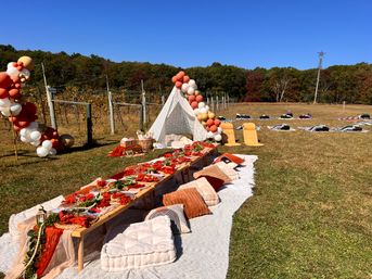 Boho vineyard picnic setup with a low wooden table on blankets, plush pillows, autumn-toned table settings and charcuterie, a lace teepee framed by peach-and-cream balloon garlands, grapevines and colorful fall trees under a bright blue sky.