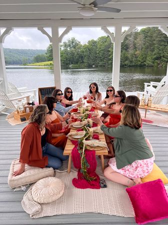 Lakefront gazebo picnic: group of friends in summer outfits toasting rosé around a low wooden table with pink decor on a covered dock by a tree-lined lake.