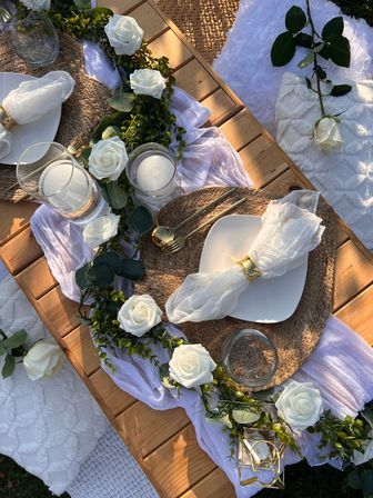 Romantic boho garden picnic on a low wooden table with white rose garland, woven placemats, gold cutlery, glass candles, airy white linen napkins and cozy blankets