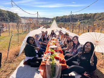 Group of people enjoying a long outdoor picnic in a vineyard during autumn — parasols, sunglasses, string lights and a rustic table spread with fall décor.
