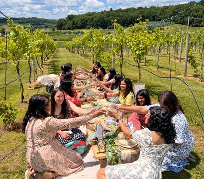 Cheerful friends toasting at a long picnic table in a sunlit vineyard, surrounded by neat rows of grapevines, rolling green hills and a bright blue sky — summer wine-tasting countryside scene.