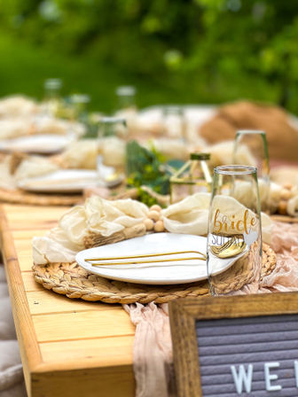 Boho outdoor wedding tablescape on a wooden table with woven placemat, white plate, gold cutlery, neutral linens and a glass labeled 'bride squad' against a green garden backdrop.