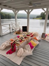 Boho lakeside bachelorette picnic under a white gazebo on a wooden dock, low wooden table with place settings, plush floor cushions and vibrant pink and yellow accents overlooking a misty, tree-lined lake.