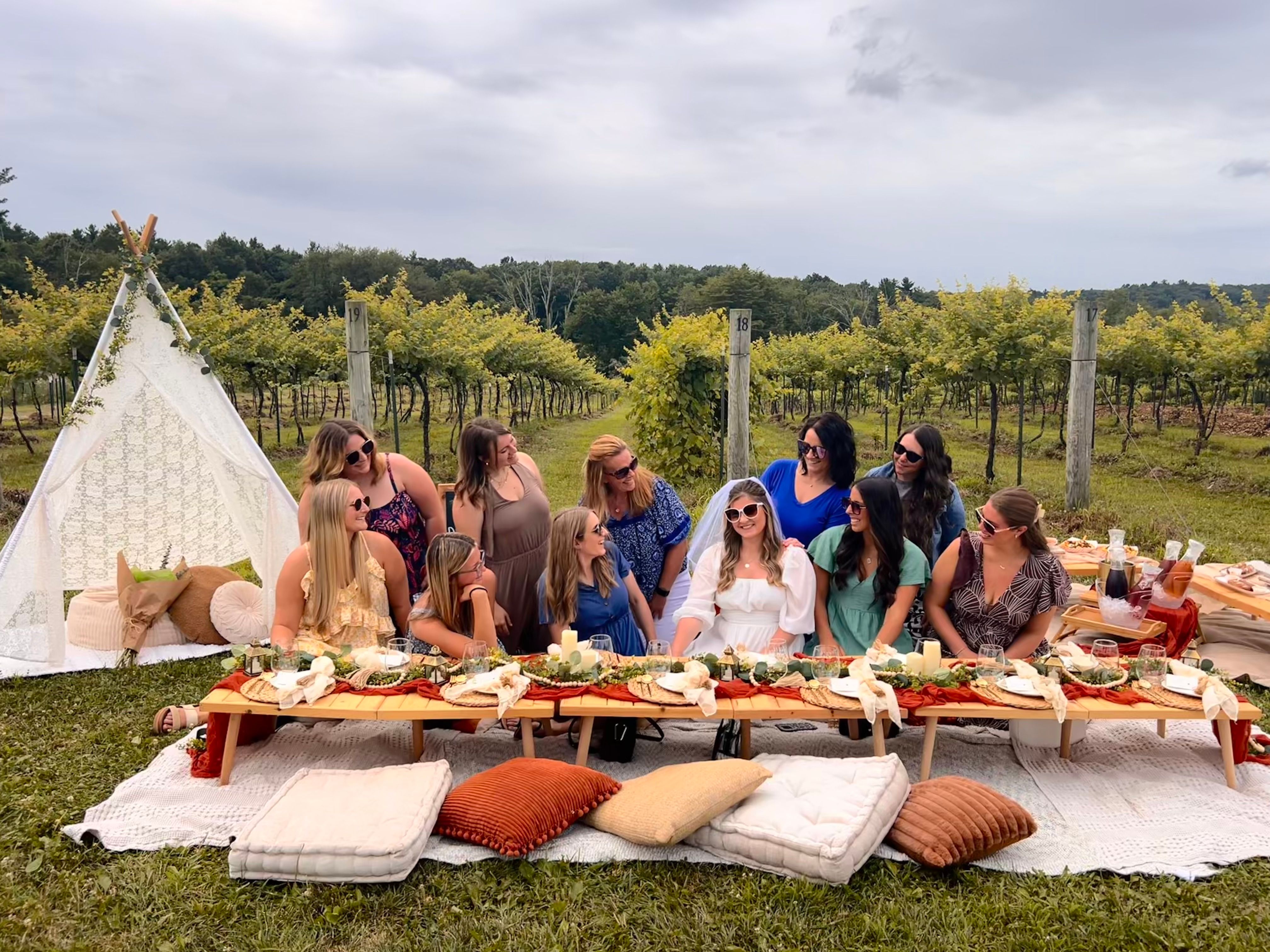 Friends enjoying a boho vineyard picnic with a low wooden table, cushions, lace teepee, rustic place settings, and a bride-to-be in a white veil among rows of grapevines under an overcast sky.