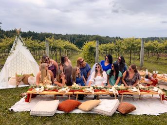 Friends enjoying a boho vineyard picnic with a low wooden table, cushions, lace teepee, rustic place settings, and a bride-to-be in a white veil among rows of grapevines under an overcast sky.