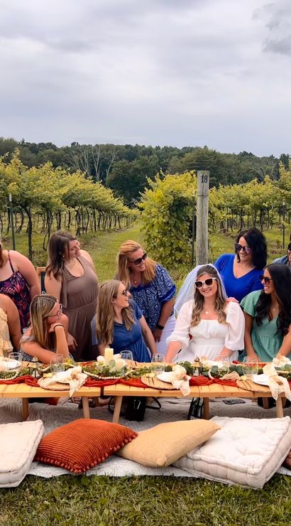 Friends enjoying a boho vineyard picnic with a low wooden table, cushions, lace teepee, rustic place settings, and a bride-to-be in a white veil among rows of grapevines under an overcast sky.