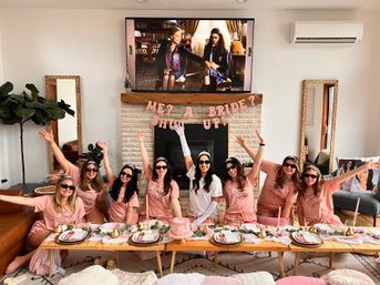 At-home bachelorette party in a cozy living room: bride-to-be in white and tiara with bridesmaids in matching pink 'Team Bride' shirts and sunglasses around a low table with pink cake, place settings and a playful banner above the fireplace.