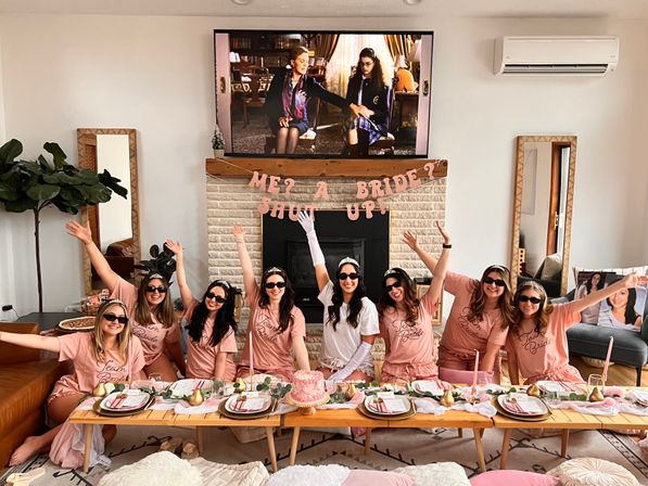 At-home bachelorette party in a cozy living room: bride-to-be in white and tiara with bridesmaids in matching pink 'Team Bride' shirts and sunglasses around a low table with pink cake, place settings and a playful banner above the fireplace.