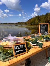 Lakeside picnic setup on a wooden table by a calm pond, styled with greenery, woven placemats, covered charcuterie and chalkboards reading “Reserved picnic” and “Girls Gone Mild” under a bright blue sky.