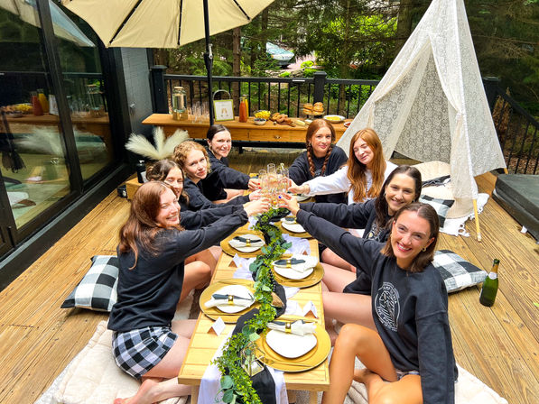 Eight friends toasting at a styled outdoor brunch on a wooden backyard deck — low picnic table with gold chargers, greenery garland, pillows, a lace teepee and patio umbrella, snacks and drinks on a side table with trees in the background.