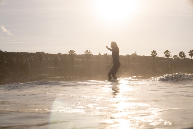 Silhouette of a surfer balancing on a longboard at sunset, golden sunlight reflecting on gentle waves with palm trees along the distant shoreline.