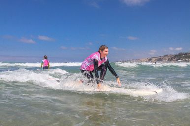 Smiling surfer in a pink rash guard kneeling on a white surfboard in shallow breaking waves at a sunny coastal beach with cliffs in the distance, beginner surf lesson vibe.