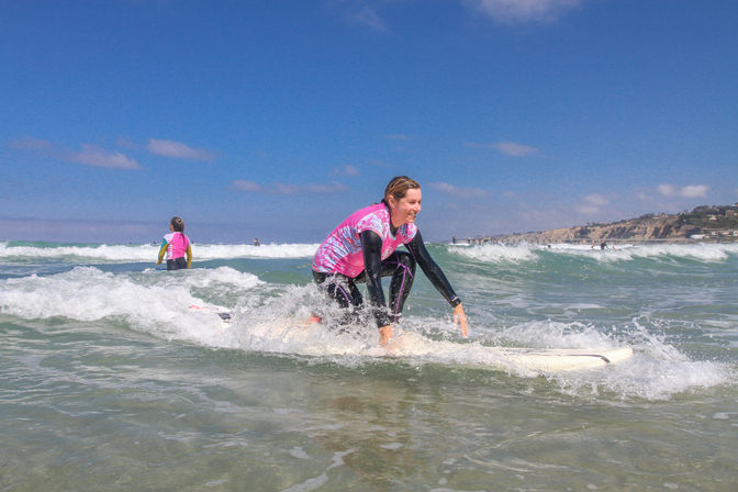 Smiling surfer in a pink rash guard kneeling on a white surfboard in shallow breaking waves at a sunny coastal beach with cliffs in the distance, beginner surf lesson vibe.