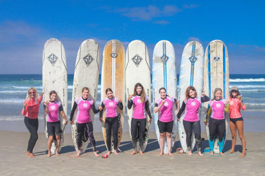 Cheerful group of women surfers in pink rash guards posing with longboard surfboards on a sunny sandy beach with blue ocean waves, striking shaka signs