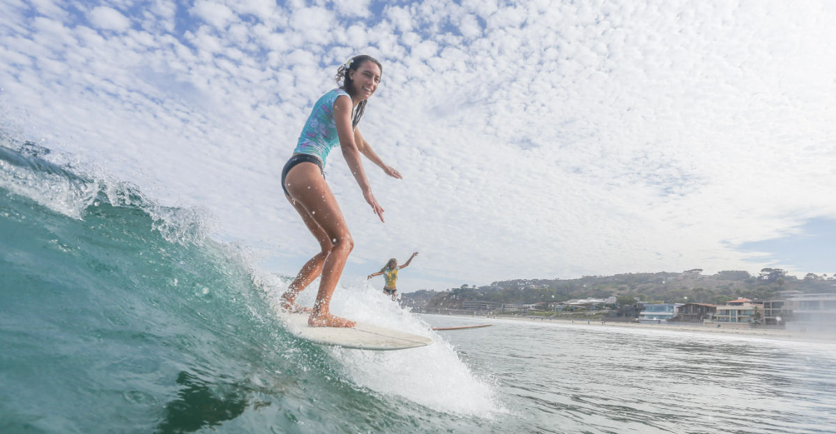Smiling woman riding a longboard on a green ocean wave near a sandy coastline with beach houses and another surfer in the background under a bright, cloud-dappled sky