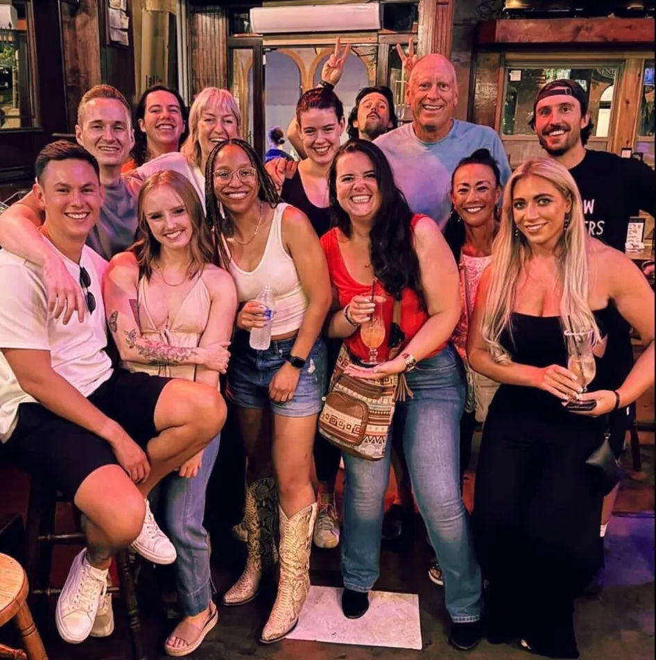Group photo of friends on a lively night out at a bar — smiling adults in casual summer outfits holding cocktails and a water bottle, one wearing cowboy boots, fun atmosphere