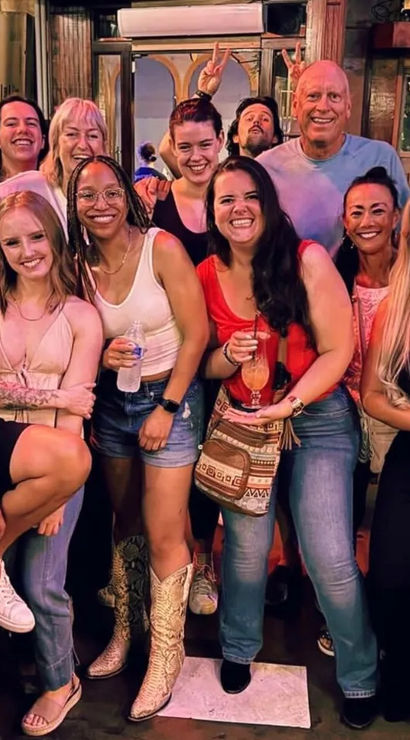 Group photo of friends on a lively night out at a bar — smiling adults in casual summer outfits holding cocktails and a water bottle, one wearing cowboy boots, fun atmosphere