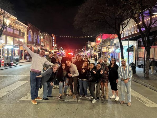 Group of friends posing in the middle of a lively downtown entertainment district at night, colorful bar and restaurant signs and string lights overhead