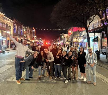 Group of friends posing in the middle of a lively downtown entertainment district at night, colorful bar and restaurant signs and string lights overhead