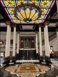Opulent hotel lobby with a large stained-glass dome chandelier, polished marble floor with geometric inlay, tall white columns with ornate capitals and a central black marble grand staircase beneath a framed portrait.