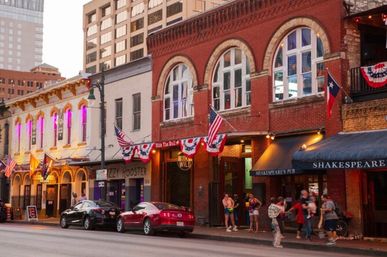 Evening on a lively downtown Texas bar street: historic brick storefronts with arched windows, festive bunting and flags, neon-lit venue fronts, parked cars and sidewalk crowds.