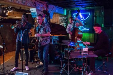 Live jazz quartet performing on a small dimly lit club stage — trumpet, saxophone, upright bass and drums under a blue neon sign.