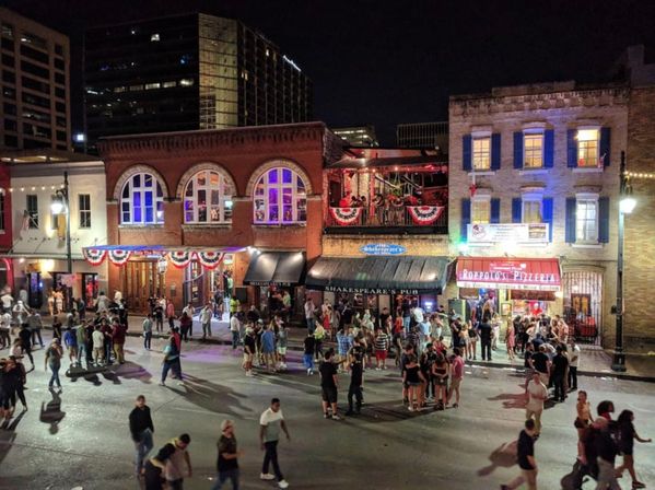 Vibrant downtown nightlife scene at night — bustling city street crowded with people in front of brick bars and restaurants with lit windows, balconies, and festive bunting.