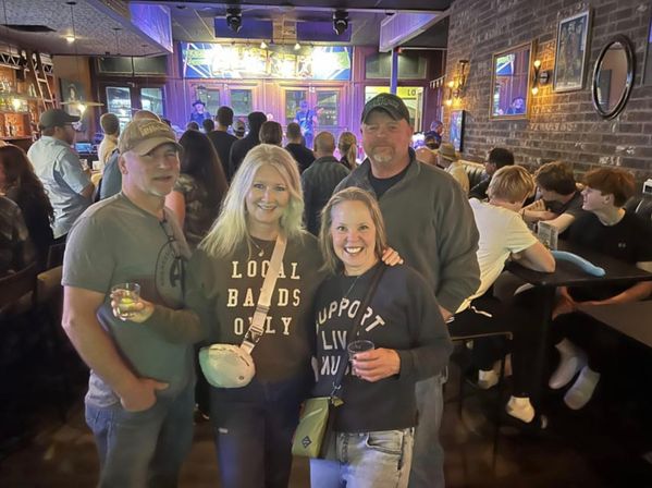 Four smiling friends pose in a packed brick-walled bar during a live band set — two women wearing sweatshirts that read “Local Bands Only” and “Support Live Music,” neon-lit stage and crowd behind them.