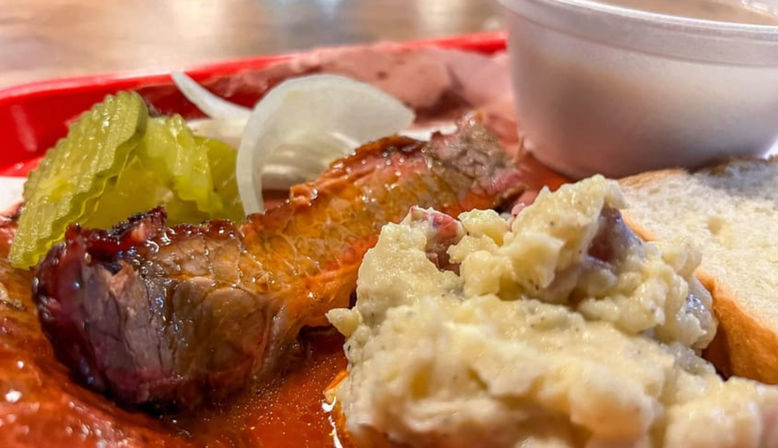 Close-up of a Southern barbecue plate: sliced smoked brisket with smoky bark and red sauce, pickle slices and sliced onion, creamy red-skin potato salad, bread and cup of sauce on a red tray