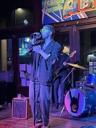 Lead singer in neon green sunglasses and oversized blazer sings into a microphone on a neon-lit bar stage with bassist and drum kit featuring a hand-horn logo — live indoor music performance.