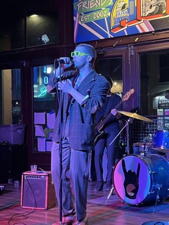 Lead singer in neon green sunglasses and oversized blazer sings into a microphone on a neon-lit bar stage with bassist and drum kit featuring a hand-horn logo — live indoor music performance.