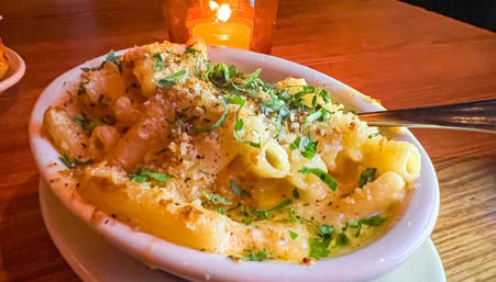 Creamy baked macaroni and cheese in a white oval dish with golden breadcrumb crust and chopped parsley, served with a spoon on a wooden table under warm candlelight.