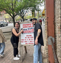 Two smiling friends pointing at a sidewalk BBQ restaurant sandwich-board sign outside a brick storefront on a tree-lined downtown street.