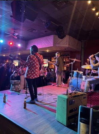 Live band playing on a small indoor bar stage — guitarist in a red heart-pattern jacket, drummer, and bassist under colorful lights with a lively crowd watching.